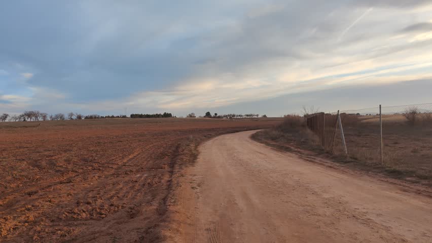 Panoramic view moving across a vast plowed field of reddish-brown soil next to a winding dirt road, revealing the rural countryside landscape under a cloudy sky during a beautiful sunset