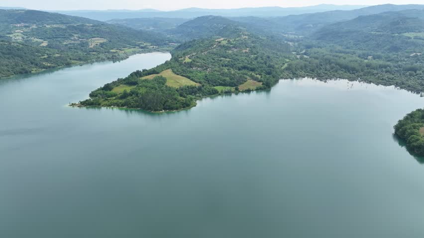 Turquoise water of Butoniga reservoir reflects white clouds in Istria region. Drone flies over artificial lake surrounded by dense green forests and rolling hills near Motovun.
