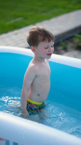 Cute toddler playing and splashing water in small inflatable pool in backyard on sunny summer day. Happy childhood, outdoor summer leisure, family home lifestyle, natural light, carefree moment.