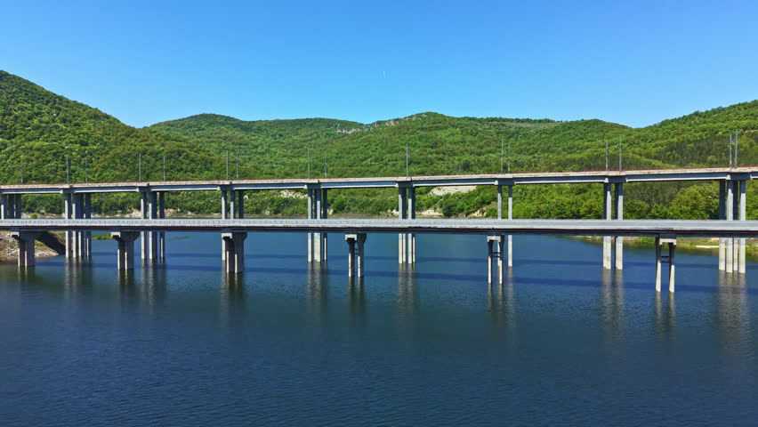 A bridge stretches over a body of water surrounded by green hills and trees. The sun shines brightly in the clear blue sky. The scene captures the structure and natural beauty of the area.
