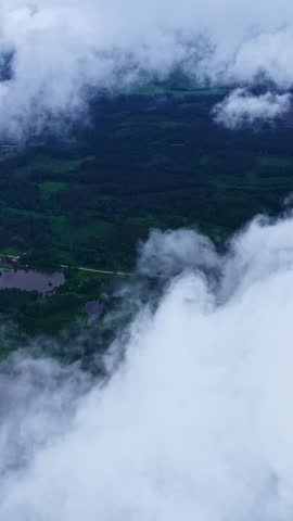 Aerial overview peeking through drifting clouds over forested countryside with lakes below, drone vertical background