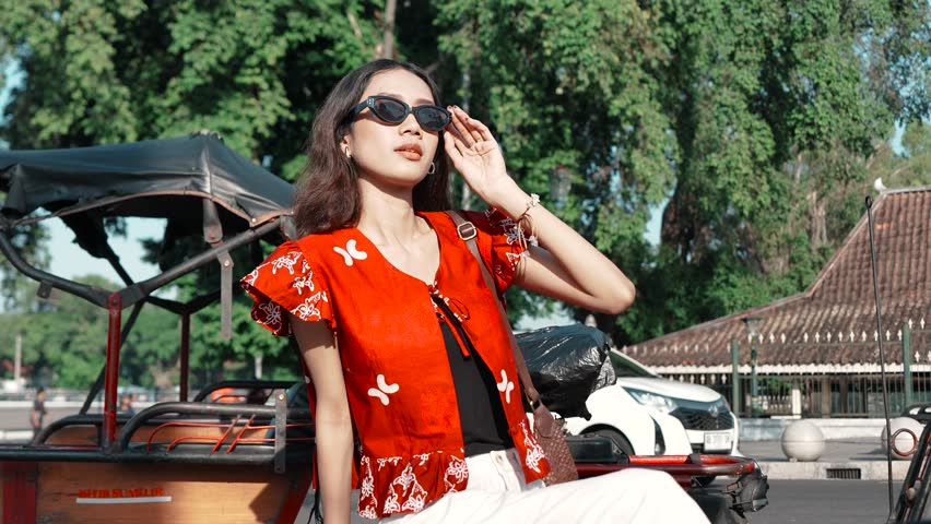 Confident young Asian woman wearing Indonesian traditional batik shirt is sitting and posing to the camera.