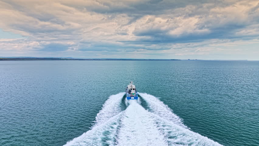 A fishing boat travels across the water in a coastal area. The scene shows waves and clouds above during late afternoon. The boat leaves a white trail behind as it moves.