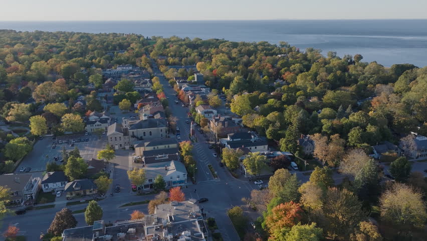 A drone flies over Queen Street in Niagara-on-the-Lake during autumn, the Memorial Clock Tower standing among the historic buildings framed by colorful fall trees.