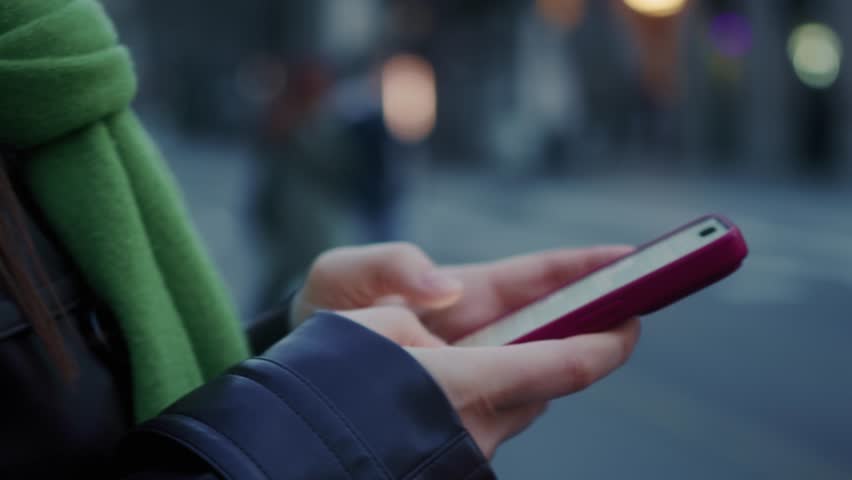Close-up of a hand holding a smartphone at a pedestrian crossing, with people walking and a busy city street in the background. High quality 4k footage