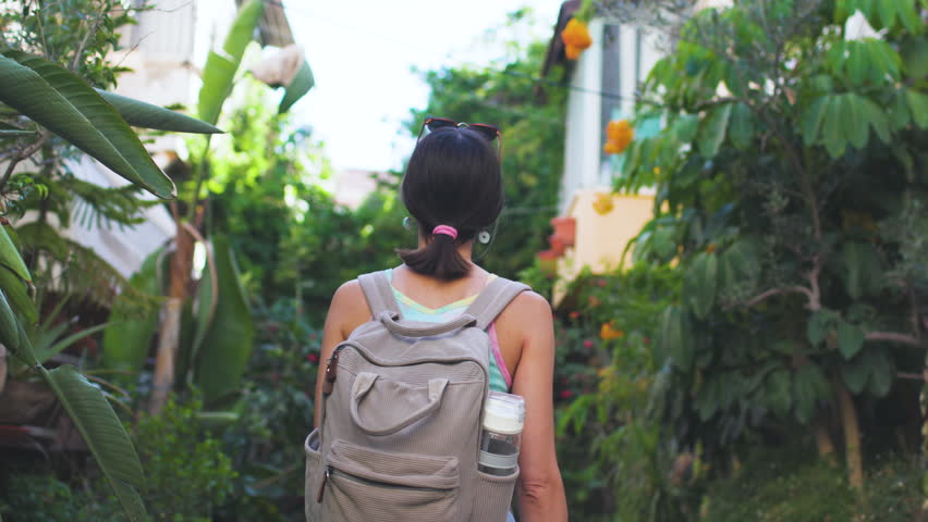 A beautiful tourist with a backpack walks along a tree-lined street, holding a phone. A young woman with a phone enjoys a stroll through the city.
