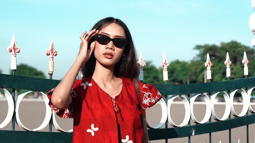 Attractive young Asian woman wearing Indonesian traditional batik shirt and sunglasses is posing in front of a gate.