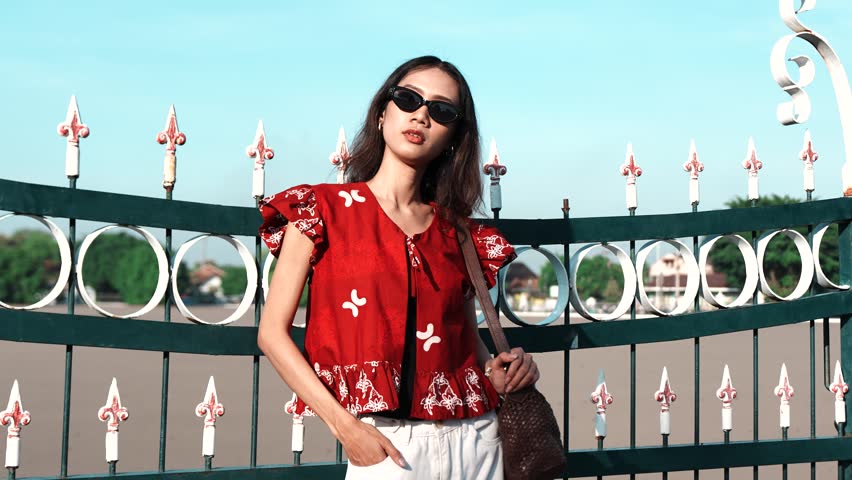 Attractive young Asian woman wearing Indonesian traditional batik shirt and sunglasses is posing in front of a gate.
