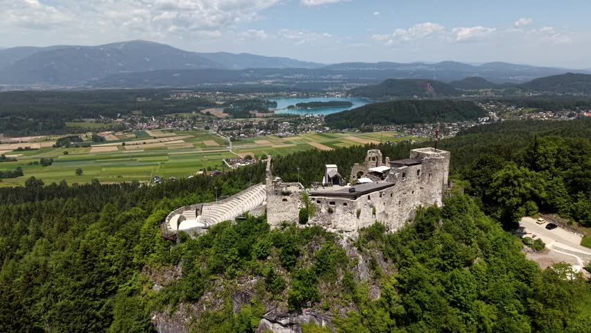 Historic Ruined Finkenstein Castle with amphitheater crowns forested hilltop above Carinthian valley, Lake Faak and Alpine peaks under cloudy skies in Carinthia, Austria. Aerial drone orbit wide shot