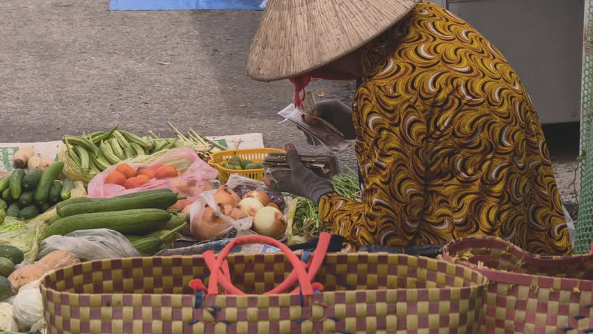 An elderly Vietnamese street vendor counting money while sitting at a traditional outdoor produce market
