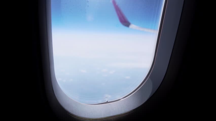 Passenger airplane cruising with oval window frame and wingtip visible outside over clouds. Cabin interior edge contrasting with bright sky and scattered clouds below. Flight experience, travel by air