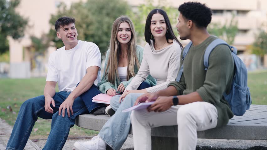 Four young multiethnic college students chatting and laughing together. Friends sitting on a bench in the university campus park having fun
