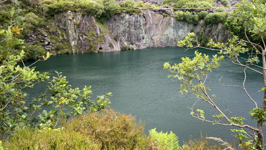 A wide-angle tilt up landscape view looking directly at the imposing sheer rock cliffs and deep turquoise water of the flooded Dorothea Slate Quarry in Talysarn, Snowdonia, North Wales, UK