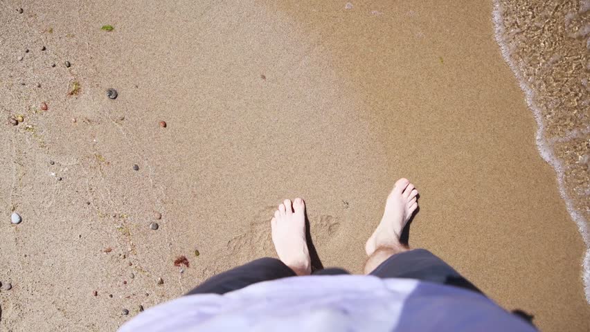 Bare feet step gradually into wet sandy shore as soft ocean waves touch skin. Warm sunlight illuminates peaceful beach environment creating soothing summer atmosphere