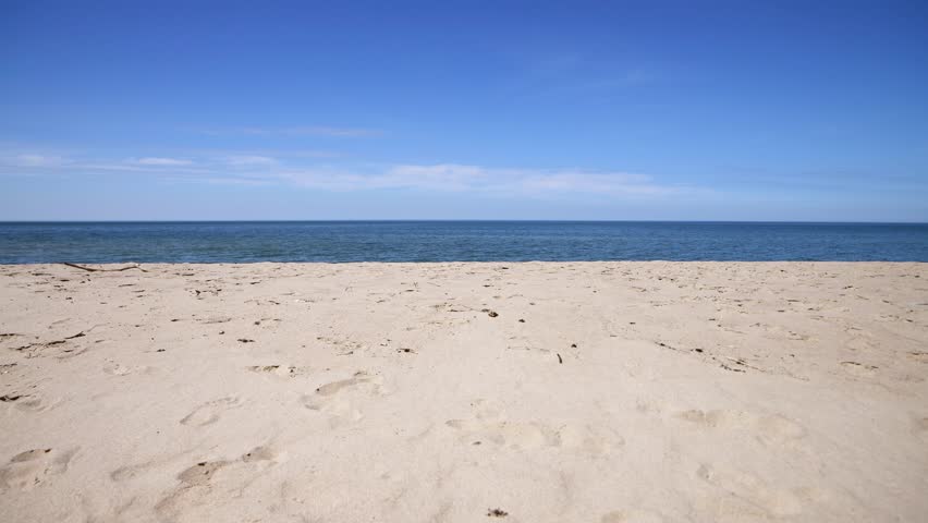 Wide empty beach with fine sand stretches toward calm blue sea under bright daylight. Horizon line separates vibrant ocean water from clear sky with minimal clouds on peaceful coastal scene