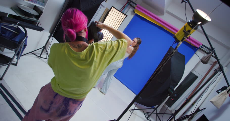 A female photographer with pink hair takes photos of a female model in a studio with a blue backdrop. The model poses while the photographer adjusts lighting and camera settings.