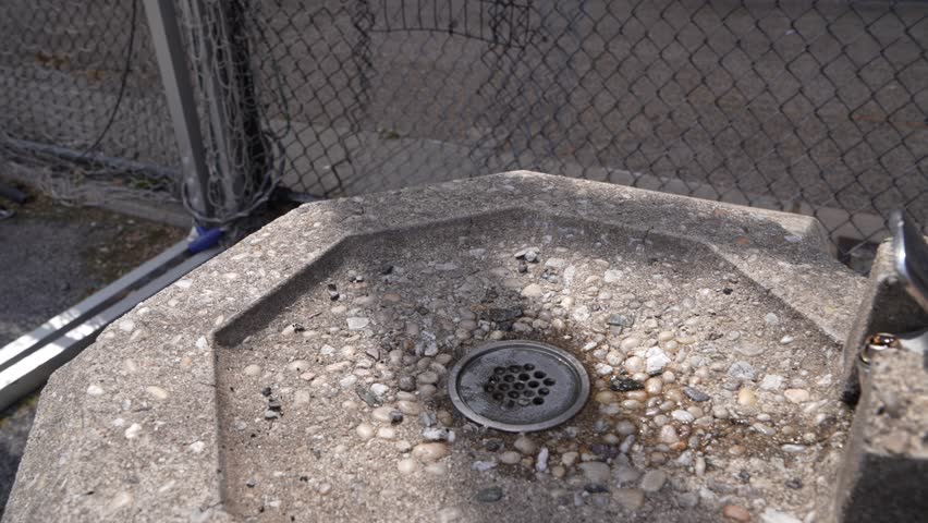 Close-up video of an old, gritty drinking fountain with a chain link fence in the background.
