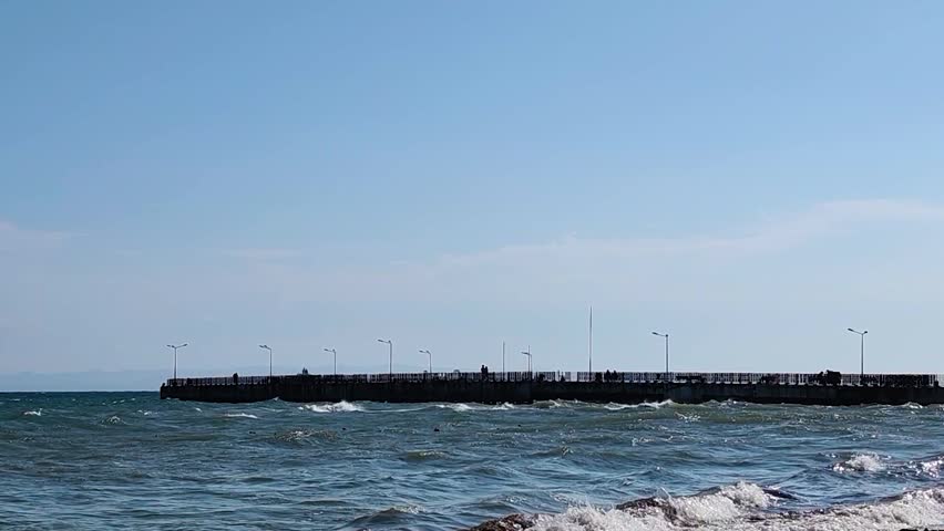 A wide-angle view of a long concrete pier stretching across a deep blue, windy sea. Choppy waves roll and create white foam in the foreground, while a few distant silhouettes of people stroll along the dock equipped with street lights. The horizon shows faint mountains under a clear sky, capturing a breezy summer day at the coast. Great for travel, maritime, seasonal tourism, and coastal architecture.