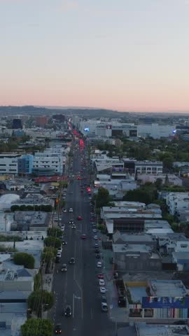 Forwards fly above wide street and buildings in urban neighbourhood at dusk.