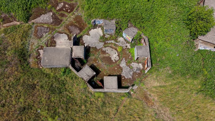 Chadwell Heath ZE1 gun emplacements seen from above as drone reverses and rises, revealing octagonal concrete AA battery remains, overgrown grassland, WWII heritage landmark in London, UK.
