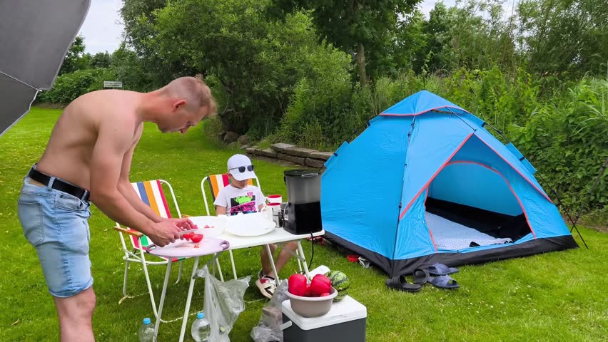 Family camping in nature, father slicing watermelon at picnic table while son sits nearby, blue tent on green grass.