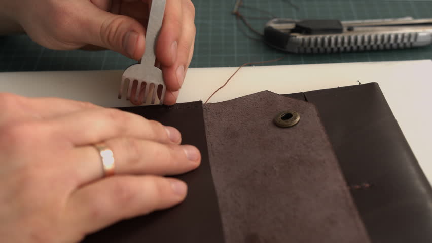 Craftsman perforating brown leather wallet with pricking iron tool for hand stitching in a traditional workshop closeup
