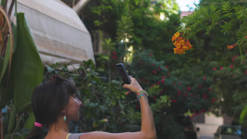 A smiling woman takes a photo of pink flowers on a smartphone on a sunny day. Social media content creation. Travel and vacation.