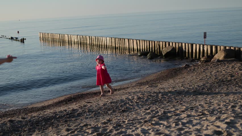Little girl in pink dress running towards her mother on sandy beach. Child playing with woman near sea water. Family activity during vacation on summer shore.
