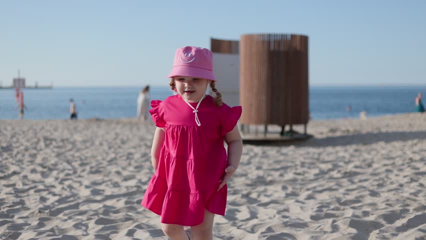 Little girl in pink dress and bucket hat walking on sandy beach. Child playing outdoors by sea during summer vacation. Happy toddler enjoying sunny day at shore.