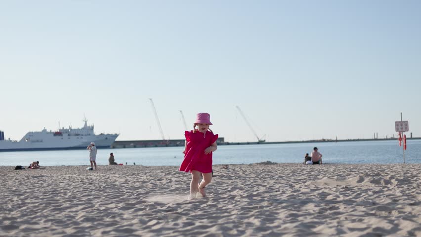 Little girl in pink dress and hat running along sand on beach. Child playing outdoors near sea during summer vacation. Leisure recreation at seaside, activity.