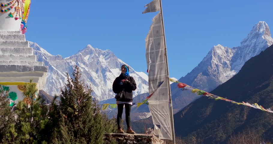 Namche Bazaar, Nepal, drone pull-out shot of a trekker standing beside a traditional Buddhist stupa as people walk along a muddy mountain slope beneath Mount Everest and Lhotse and green hills