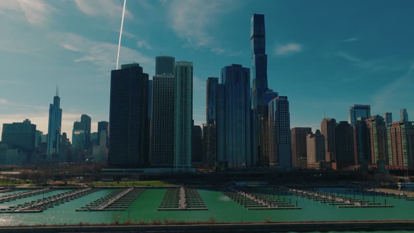 Aerial of an empty marina harbor on Lake Michigan with rows of boat docks and finger piers, a moored ship and the Chicago skyline beyond under a clear blue spring sky.
