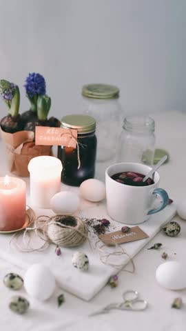 A person stirring a cup of tea with a spoon surrounded by easter eggs, flowers, and candles on a table.