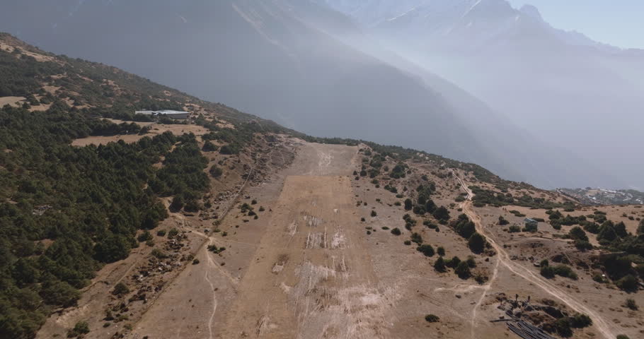 Namche Bazaar, Nepal, aerial drone wide shot overlooking a rugged high-altitude dirt clearing and mountain ridge surrounded by green hills and a hazy Everest Region valley view.