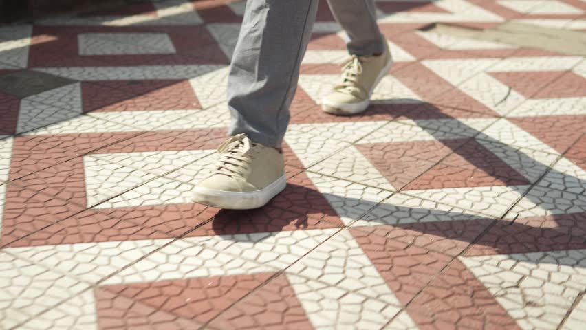 Detail close up of businessman legs in gray pants and beige sneakers walking with shadow on geometric patterned pavement
