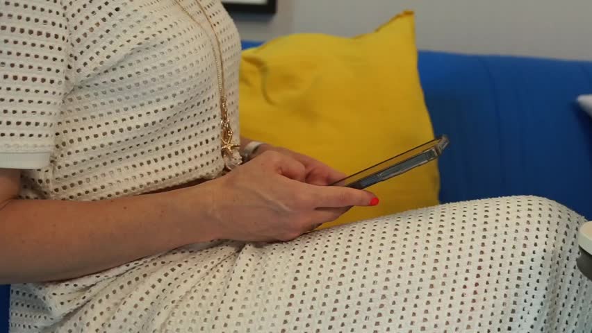 Side view of a fashionable woman wearing a white laser-cut pattern dress, sitting on a blue sofa and browsing on her mobile phone before setting it down.