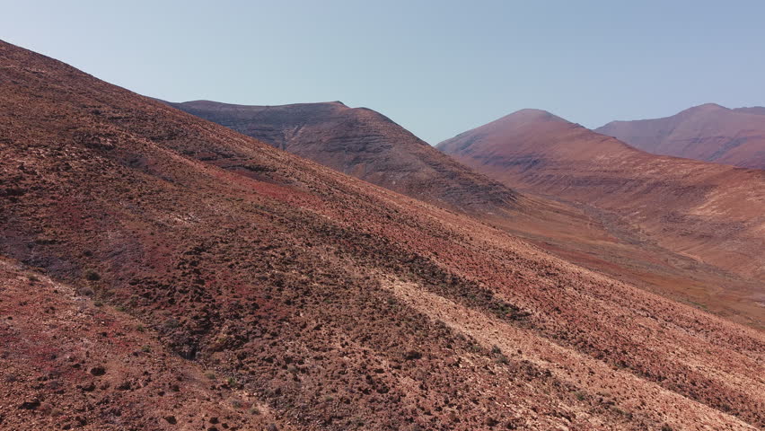 Drone footage flying above a rocky volcanic mountain slope in Fuerteventura, Canary Islands, Spain. Aerial view of arid terrain, red volcanic soil and rugged hills in the island interior.