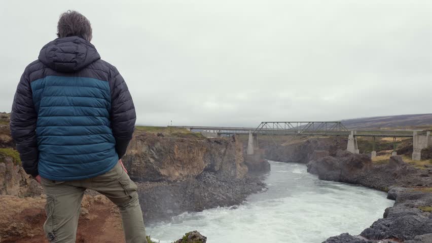 Mature man standing on a cliff admiring the majestic Godafoss waterfall in Iceland with a bridge in the background