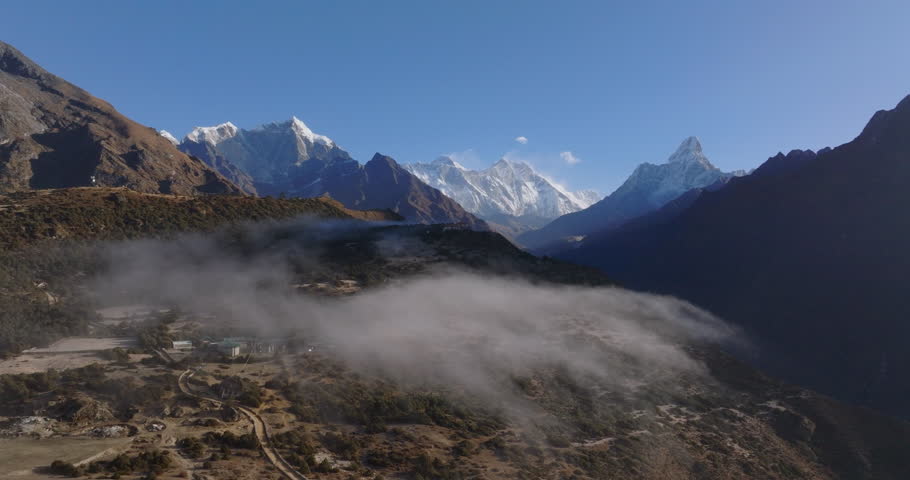 Everest region, Nepal, slow pull-out drone shot revealing a dramatic mountain valley blanketed in low-hanging fog and mist, with rugged terrain, winding trails, and snow-capped peaks of the Himalayas