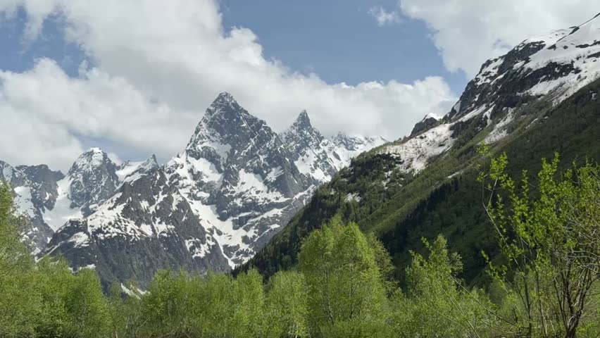 Snow-covered mountains tower over vibrant green foliage, showcasing a stunning natural landscape with a clear blue sky and scattered clouds