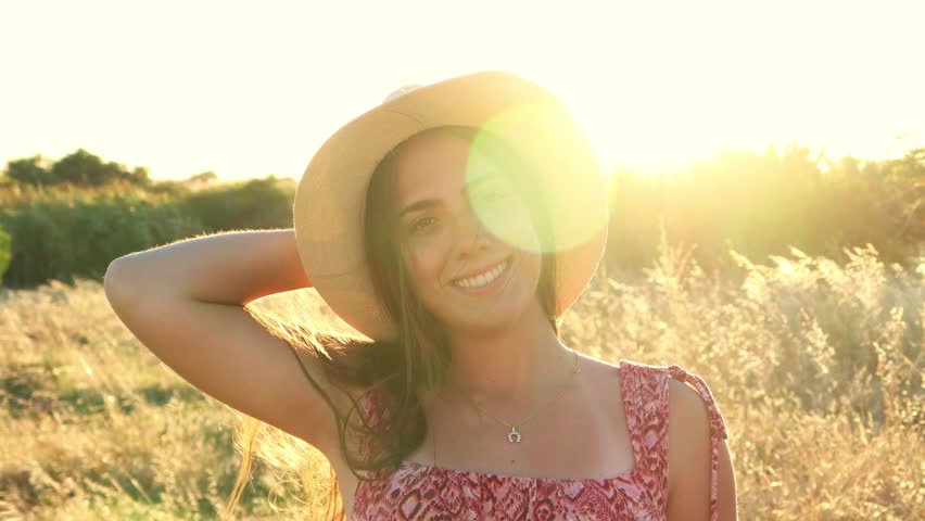 A young woman in a straw hat smiles happily, enjoying the golden hour light in a peaceful grass field. Her relaxed demeanor captures a joyful and serene outdoor portrait, celebrating nature