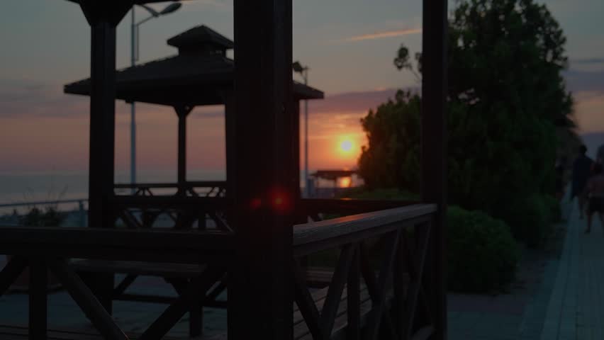 A wooden gazebo overlooking the ocean at sunset beside a paved walkway lined with bushes and walking people.
