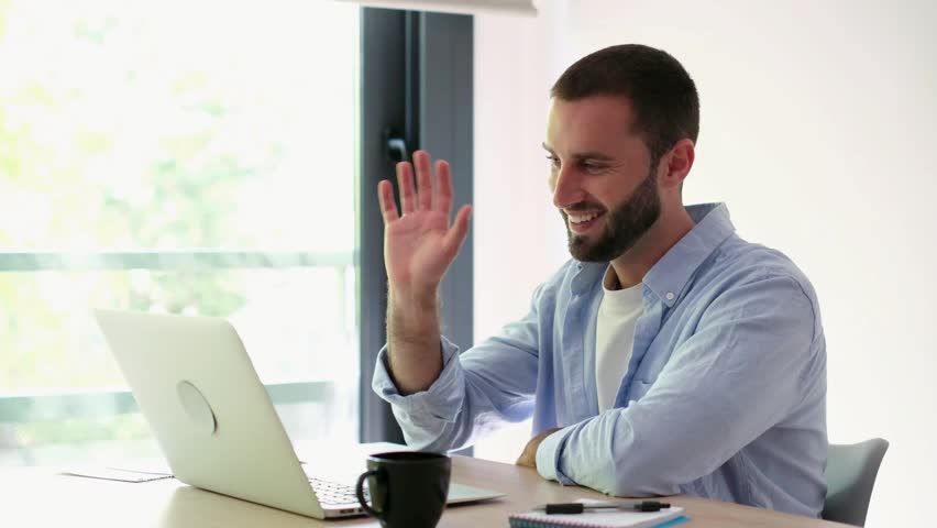 A cheerful young man is seen engaging in a video call on his laptop. He smiles, waves, talks, and gives a thumbs-up, showcasing positive and friendly online communication from his home or office.