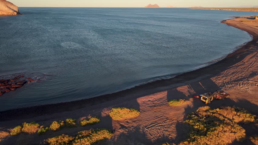 Camping on the beach in Baja California at sunset with an SUV and a teardrop camper trailer near the water and sand.