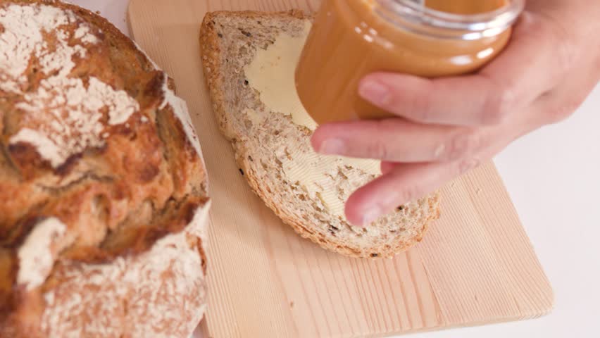 Close-up of hands spreading peanut butter on bread amidst sourdough, baguettes, and croissants.