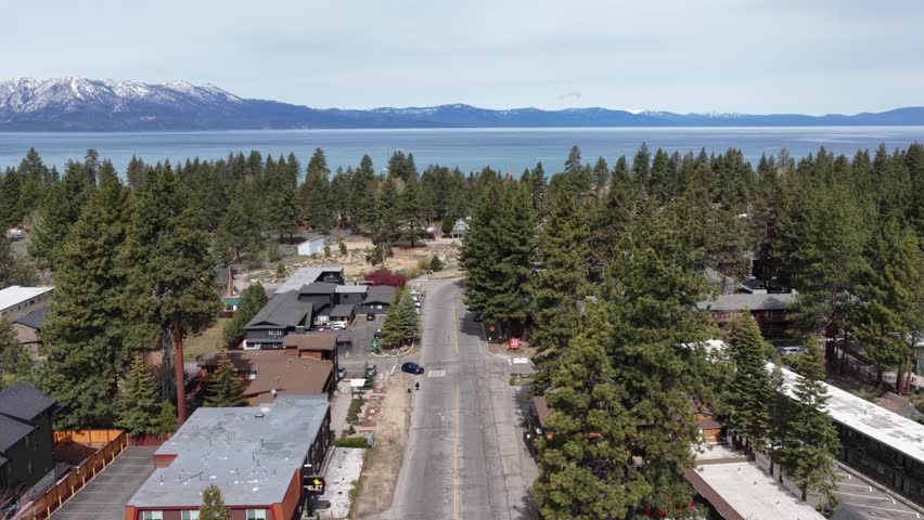 Quiet road winds through dense pine forest past alpine cabins and lodges toward turquoise Lake Tahoe with snow-capped Sierra Nevada peaks rising beyond, California, USA. Aerial drone wide shots