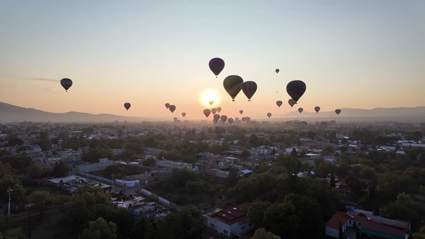POV aerial view from a hot air balloon at sunrise above Teotihuacan with dozens of colorful balloons floating over the valley and surrounding mountains in Mexico.