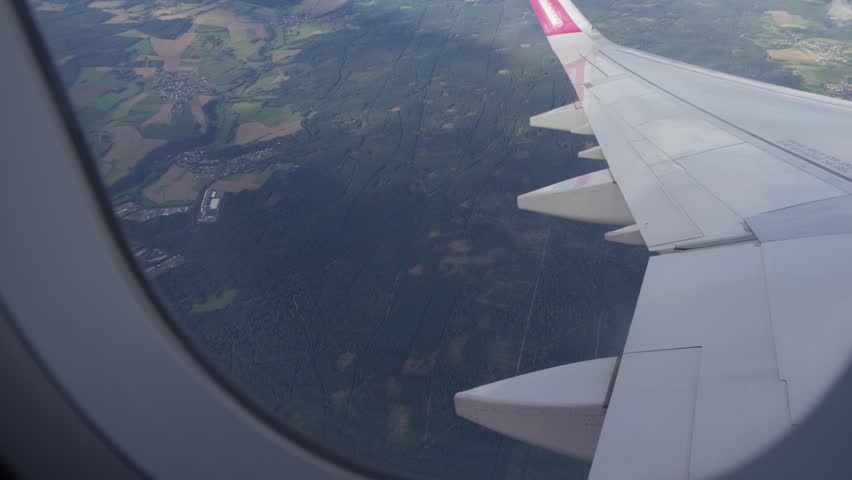 Looking through a plane window at a white wing flying over green forests in Germany.