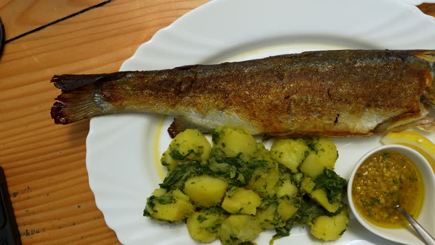 Trout is served whole with potatoes, lemon and sauce on a white plate, top-down shot