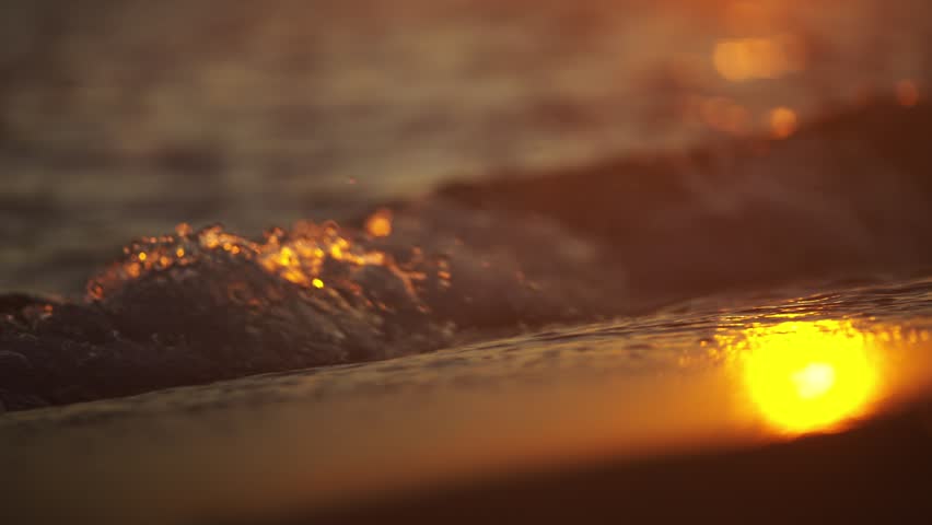 Waves washing sandy beach during sunset, golden hour light reflections.